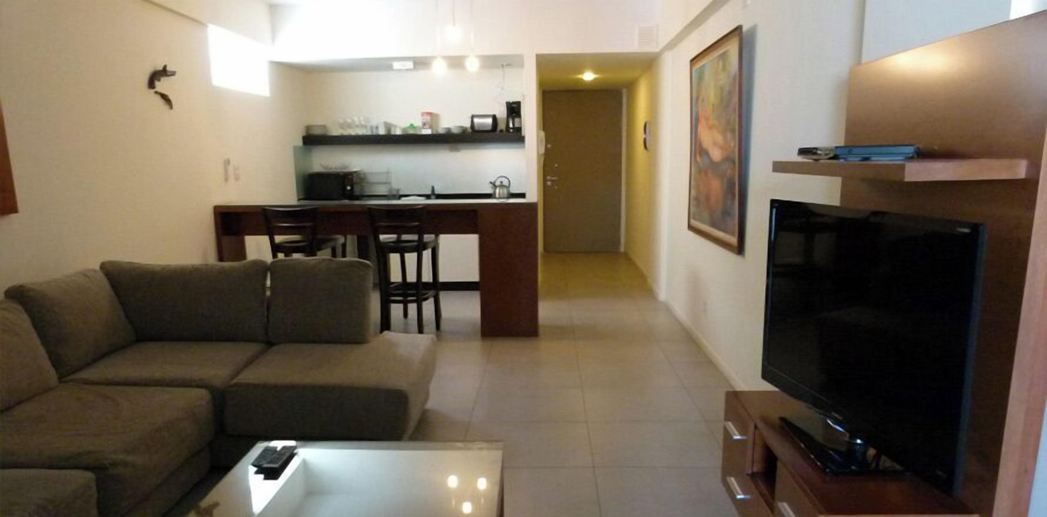 Una sala de estar con sofá, una mesa de centro, televisor y una cocina al fondo.