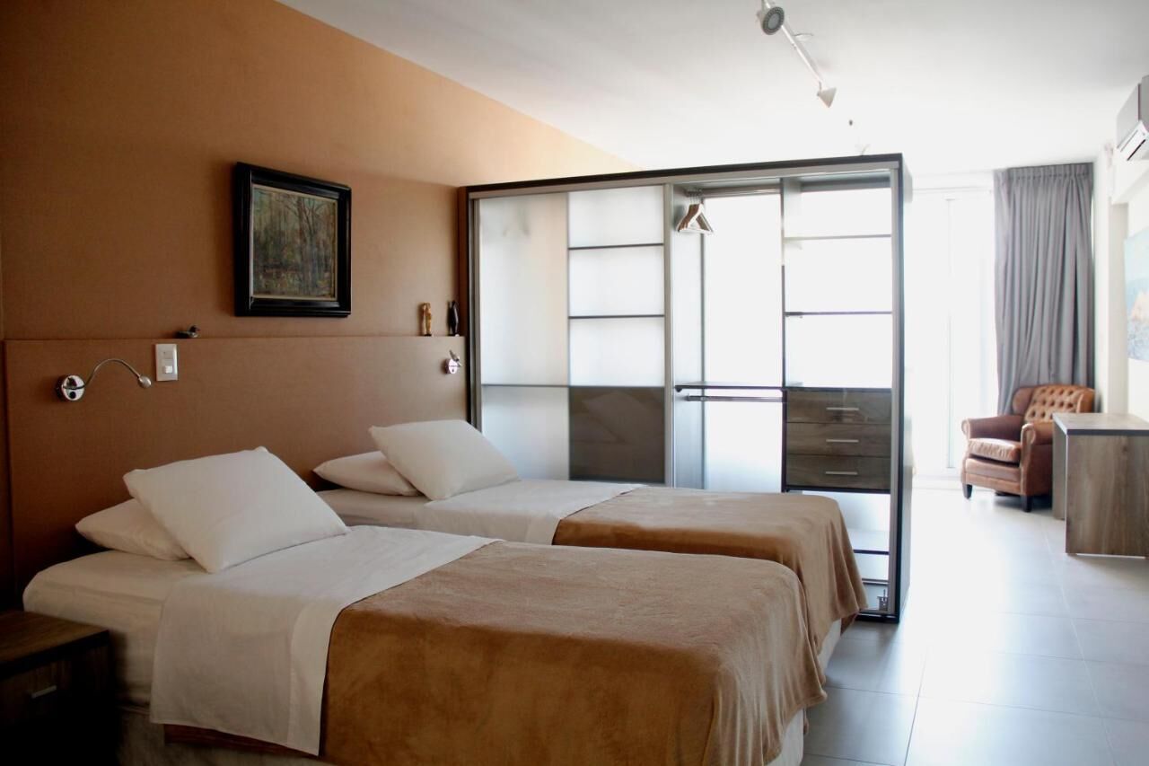 A hotel room with two beds, a sliding glass door to the balcony, a painting on the wall, and a small seating area with a chair and table.