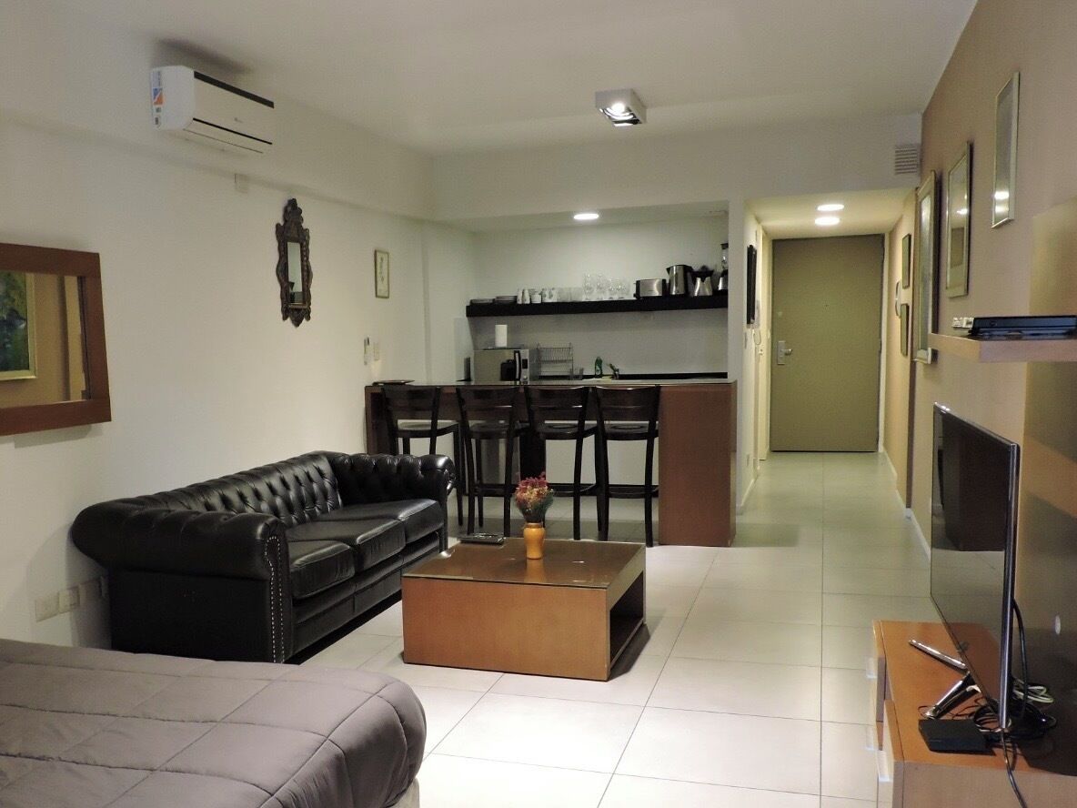 Una sala de estar con un sofá de cuero negro, una mesa de centro de madera y un sofá gris. Se observa una cocina con una mesa de comedor y sillas al fondo.