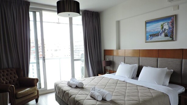A hotel room with a large bed, a chair, a nightstand, a lamp, and a painting on the wall. - Rivadavia Apartments (Buenos Aires)