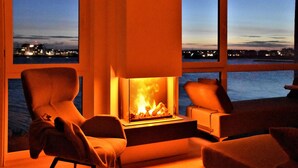 Un salon moderne avec une cheminée, deux fauteuils et une vue sur un plan d’eau au crépuscule.
