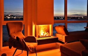 A modern living room with a fireplace, two armchairs, and a view of a body of water at dusk. - Cozy apartment in OstseeResort Olpenitz (OstseeResort Olpenitz)
