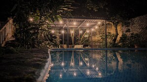 Un espace piscine aménagé pour la nuit, avec un coin salon abrité et une végétation luxuriante tout autour.