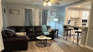 A modern living room with a dark brown leather sofa, a round coffee table, and a kitchen area with white cabinets and a bar stool.