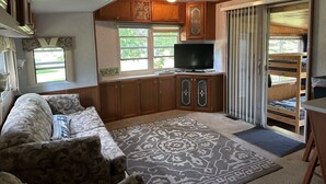 Interior compacto de autocaravana con sofá, televisión, armarios de madera y puerta corredera que da a una zona de descanso.