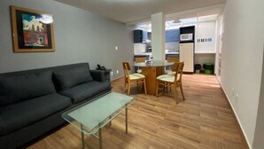 A living room with a sofa, a glass coffee table, and a dining area with chairs and a table. There is a kitchen in the background with cabinets and appliances.