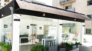 Un café moderne en plein air, doté de murs en verre, de plantes en pot et de sièges extérieurs.