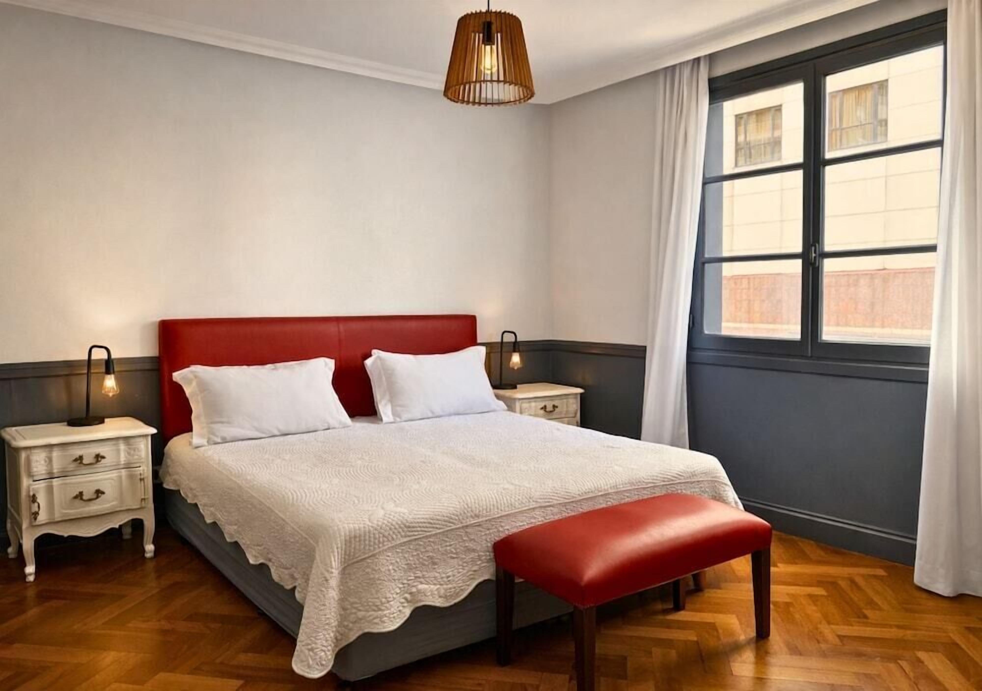 A bedroom with a red headboard, white bedding, a wooden bench, a bedside lamp, and a window with curtains.