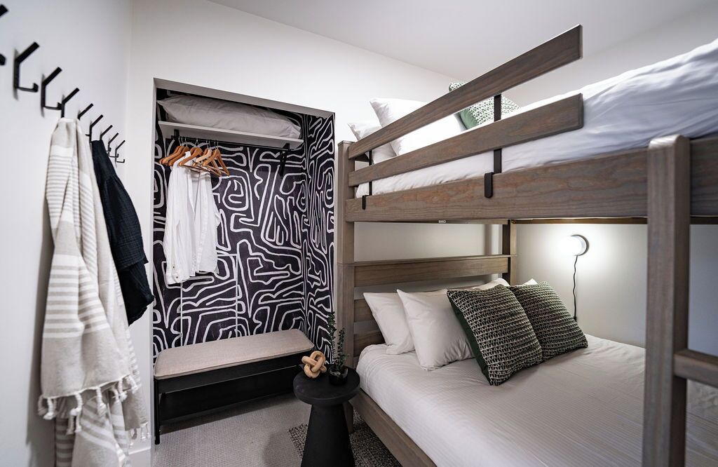 A bedroom with a bunk bed, a bedside table with a lamp, a hanging coat rack, and a patterned wall.
