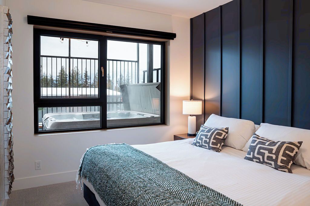 A hotel room with a large bed, a window with a view of a hot tub and trees, a nightstand with a lamp, and a dark headboard.