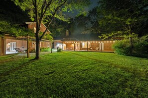 A well-lit wooden cabin with a large deck, surrounded by lush greenery.