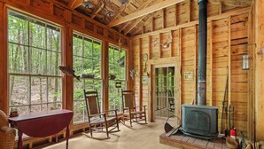 Interior de una cabaña de troncos con ventanas amplias, chimenea, mecedoras y una mesa roja.