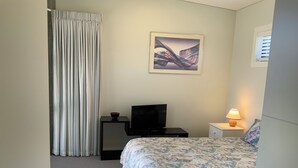 A hotel room with a bed, a television on a stand, a bedside table with a lamp, a framed picture on the wall, and a window with blinds.
