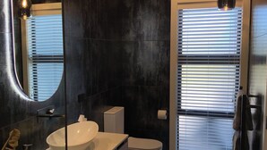 A modern bathroom with a large oval sink, a toilet, and a bidet. There is a mirror with a light, a towel rack, and a window with blinds.