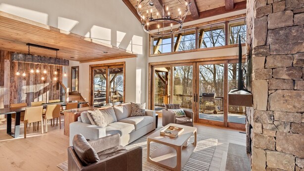 A spacious living area with a large sofa, coffee table, and dining table. There are large windows providing natural light and a view of the outdoors.