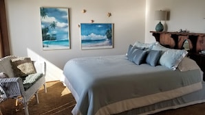A bedroom with a large bed, two wall-mounted paintings, a wooden nightstand, and a wicker chair.