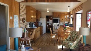 A compact kitchen with wooden cabinets, a dining area with a table and chairs, and a small seating area with a chair and a lamp.