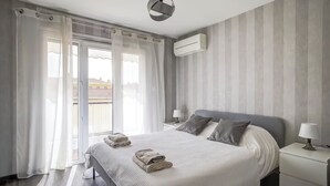 Ein modernes Hotelzimmer mit einem großen Bett, zwei Nachttischen mit Lampen, Blick auf die Stadt durch ein großes Fenster und einer Klimaanlage an der Wand.