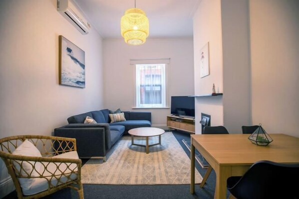 A living room with a sofa, a coffee table, a TV, and a dining table. - Beach & Pines Large 3Br House Glenelg Wifi BBQ (Glenelg South)