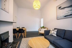 A modern living room with a fireplace, a round coffee table, a sofa, a dining area with chairs, and a painting of a house on the wall. - Beach & Pines Large 3Br House Glenelg Wifi BBQ (Glenelg South)