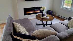 A living room with a gray sofa, a flat-screen TV mounted on the wall, a fireplace, and a window with a view of the outdoors. - Rural Town Oasis in Bonavista  (Bonavista)