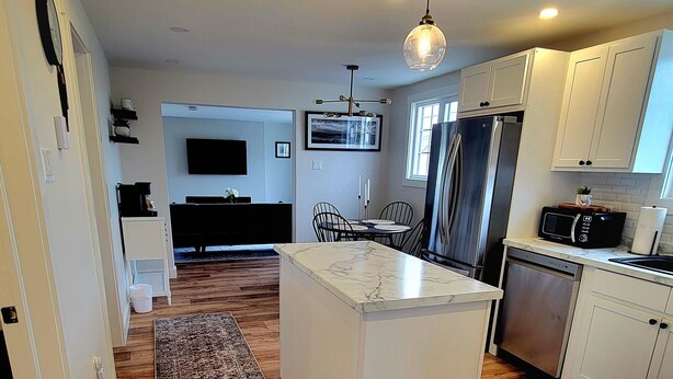 A modern kitchen with a central island, wooden flooring, and white cabinetry. - Rural Town Oasis in Bonavista  (Bonavista)