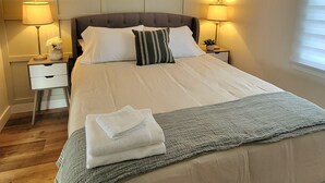 A neatly made bed with a grey blanket, two bedside lamps, a small white nightstand, and framed botanical prints on the wall. - Rural Town Oasis in Bonavista  (Bonavista)