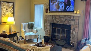 A living room with a fireplace, a television, a sofa, a striped armchair, and a lamp.