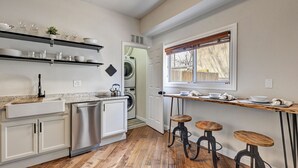 Eine Küche mit Holzboden, weißen Schränken, einer Edelstahl-Spüle, einer integrierten Waschmaschine und einem Fenster mit Jalousien. Zwei Holzhocker und ein kleiner Tisch mit Geschirr sind ebenfalls vorhanden.