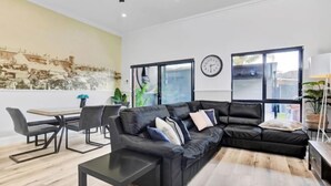 A modern living room with a black leather sofa, a dining table with chairs, a coffee table, and a large window with a view. - Noir Cottage Glenelg North 3Br Netflix BBQ (Glenelg North)