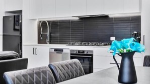 A modern kitchen with white cabinetry, a black backsplash, and a vase with blue flowers on the table. - Noir Cottage Glenelg North 3Br Netflix BBQ (Glenelg North)