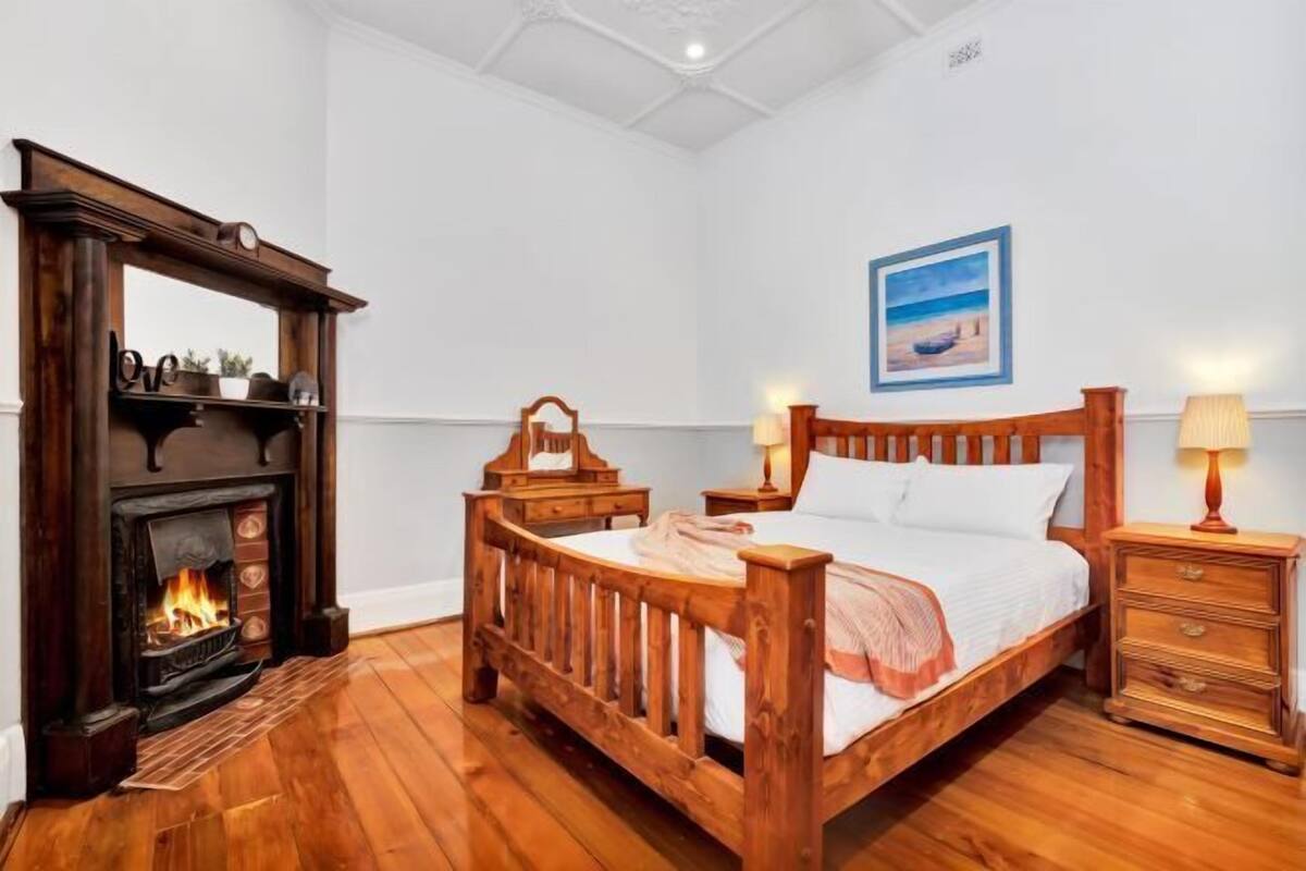 A bedroom with a wooden bed, a fireplace, a nightstand, a wall-mounted picture, and a wooden floor.