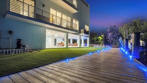 A modern two-story building with a well-lit outdoor area, a wooden deck, and a visible outdoor seating area. - Keys Casa: Elegant Waterfront Retreat, Sleeps 16, Elevator, Private Dock, Putt-Putt (Key Largo)