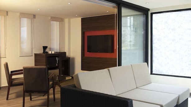 A modern living room with a sofa, a television mounted on a wooden wall, and a dining area with a table and chairs. - Mc Suites Reforma (Mexico City)