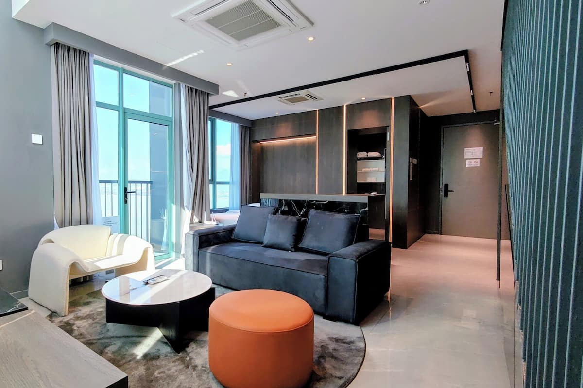 A modern hotel room with a sofa, a round coffee table, a chair, a TV, and a large window with curtains.