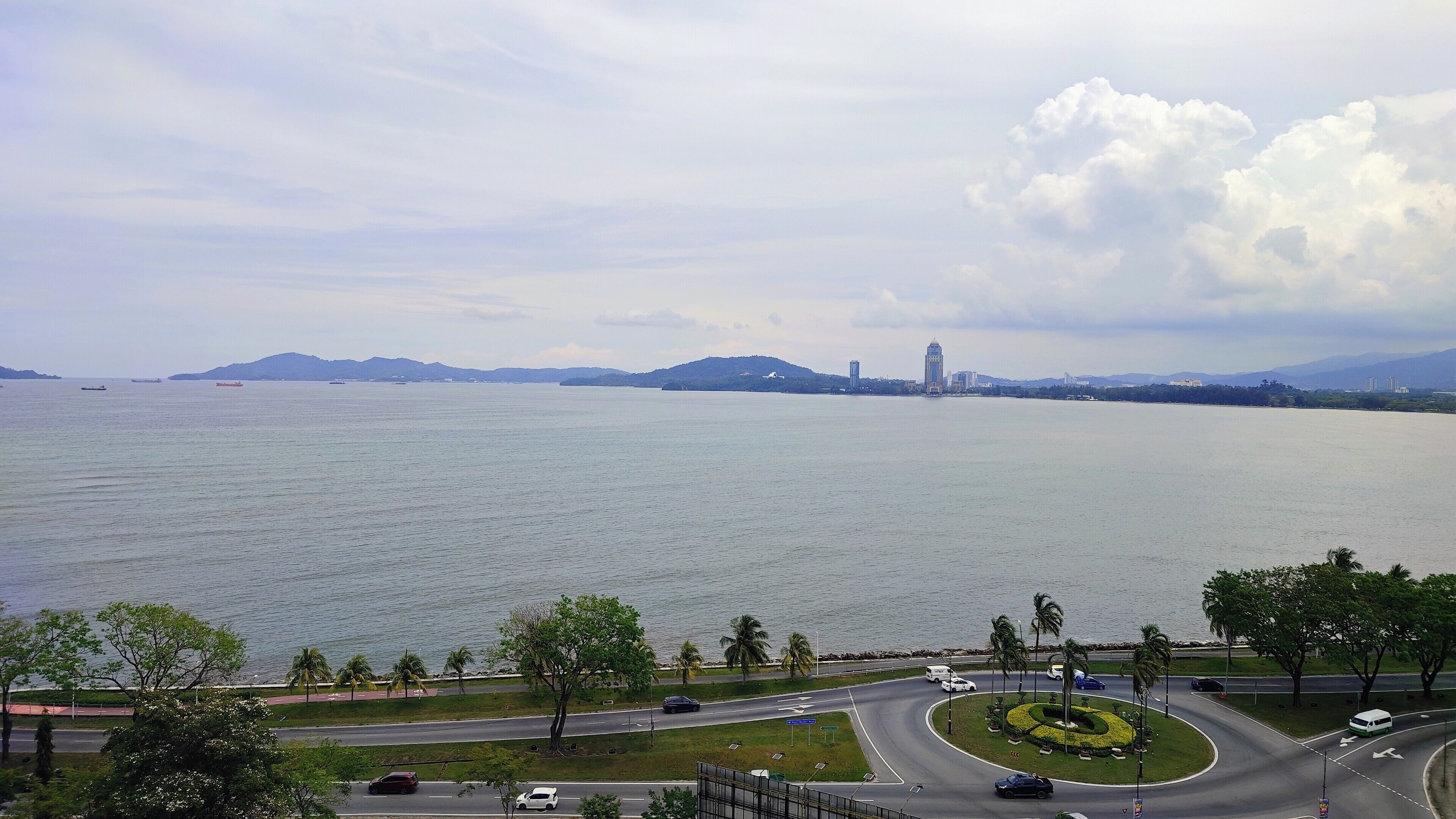 A coastal cityscape with a large body of water, a roundabout, and a prominent tower in the distance.