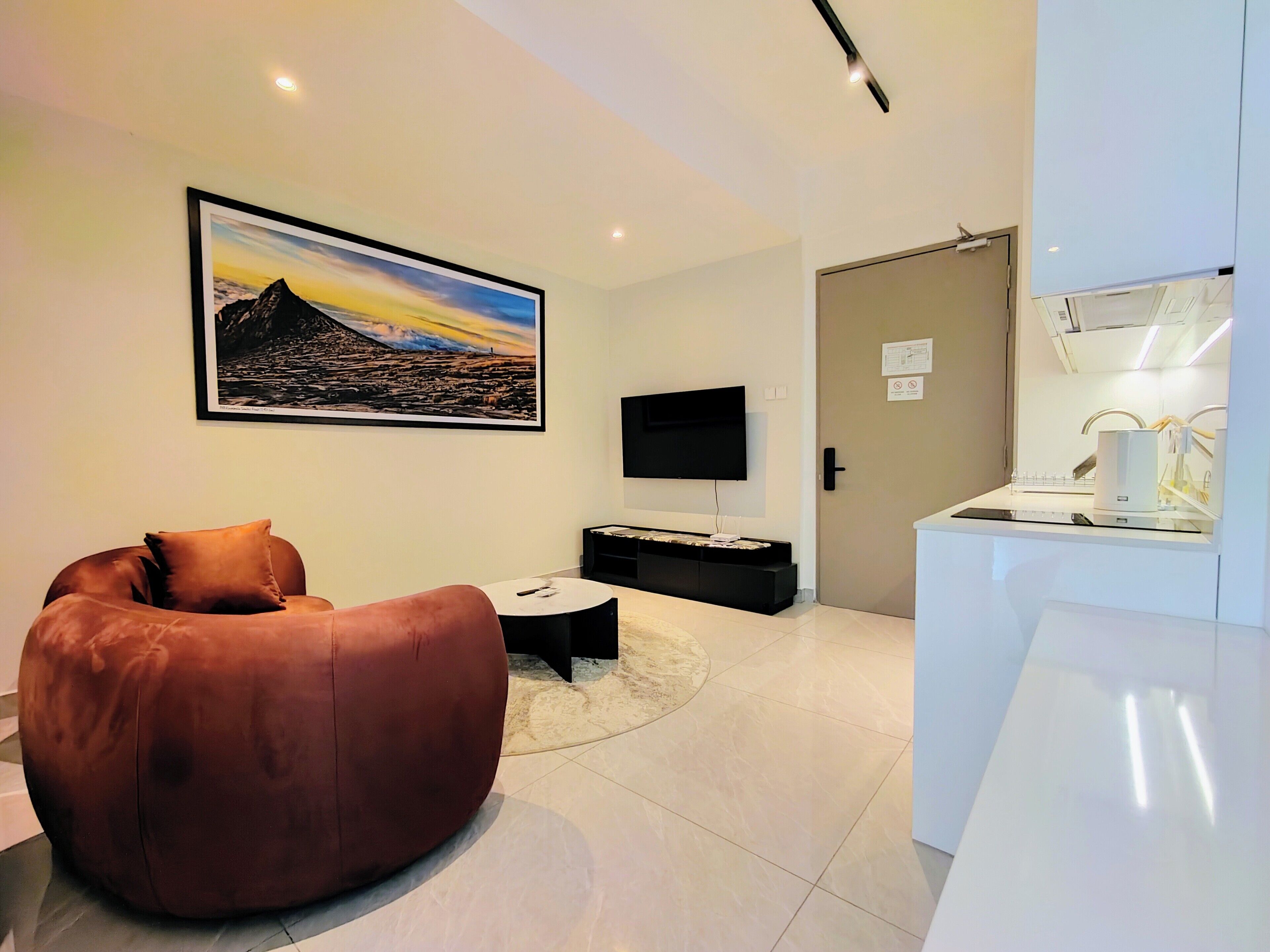 Una sala de estar moderna con un cuadro grande, un sillón informal de color marrón, un televisor de pantalla plana, una mesita de centro pequeña y una zona de cocina con encimera y electrodomésticos blancos.