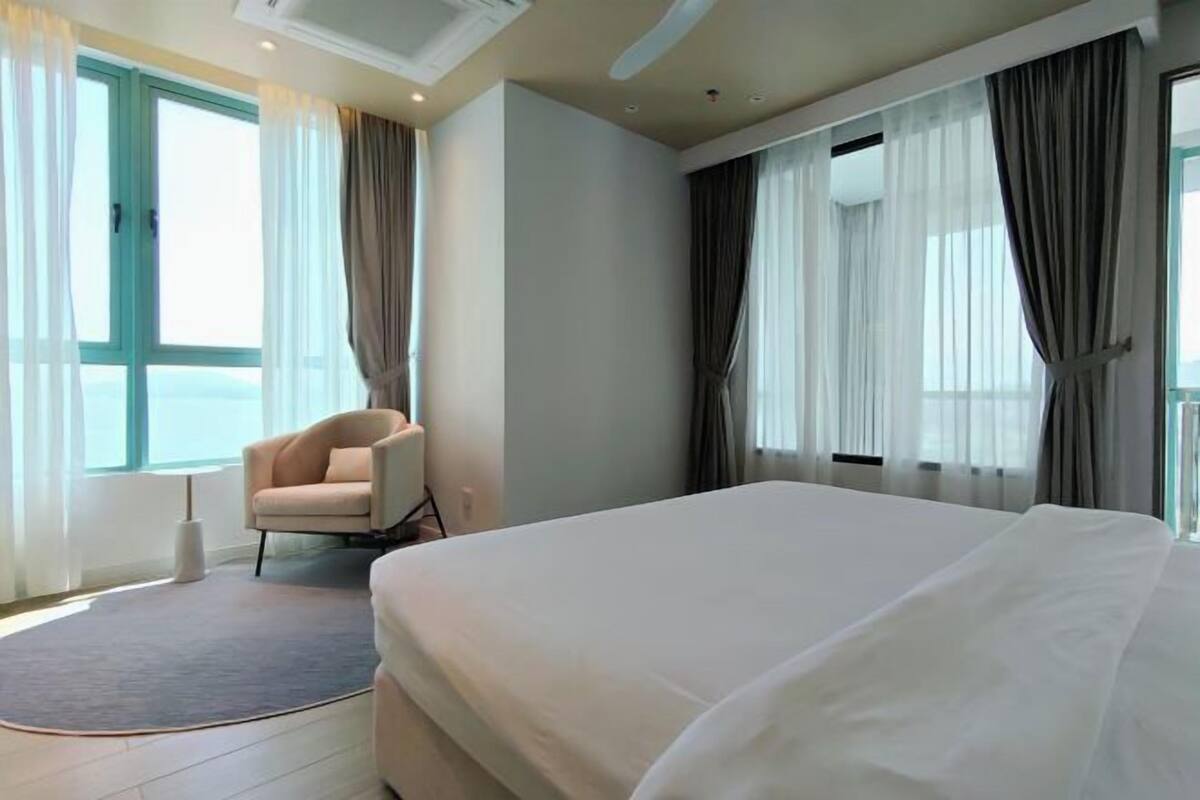 A hotel room with a bed, a chair, a window with curtains, and a view of the sea.