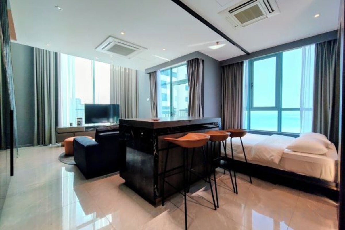 A modern hotel room with a large bed, a TV, a bar area, and a view of the city.