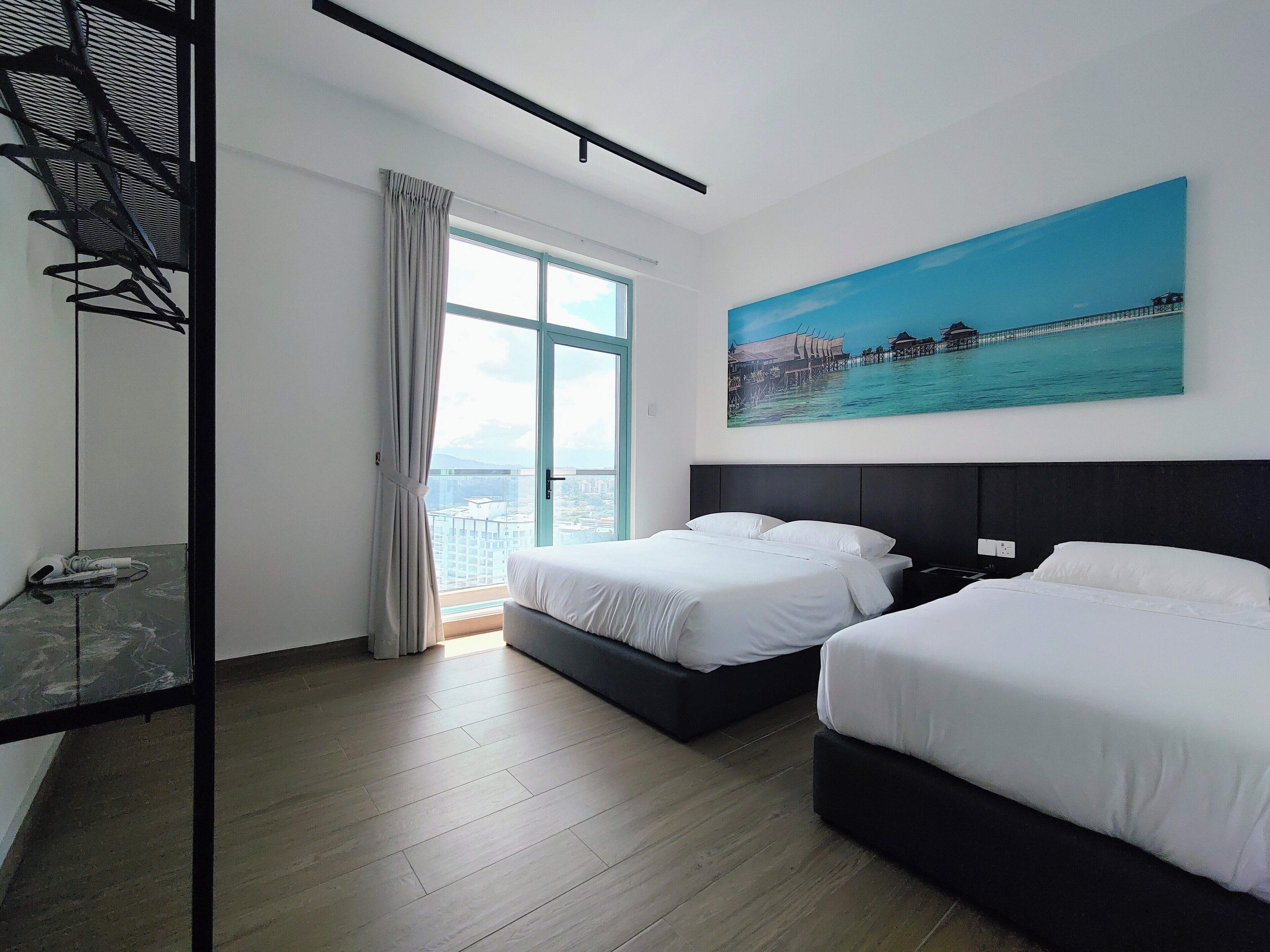A hotel room with two beds, a large window with a view of buildings, a painting of a coastal scene, and a metal shelf with hangers.