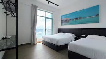 A hotel room with two beds, a large window with a view of buildings, a painting of a coastal scene, and a metal shelf with hangers.