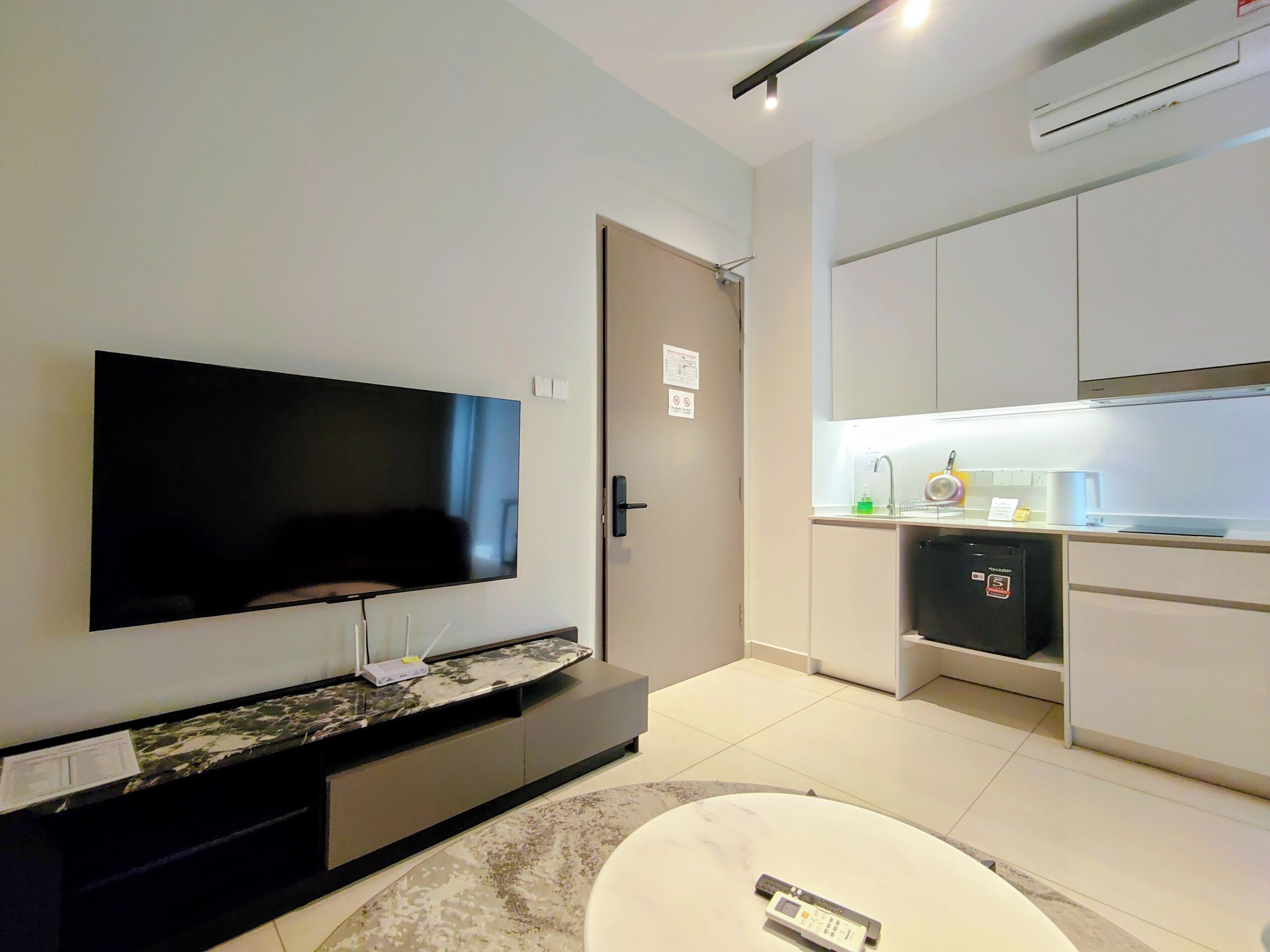 Una sala de estar moderna con un televisor de pantalla plana montado en la pared, una mesa de centro con diseño de mármol y una pequeña cocina con refrigerador y pileta.