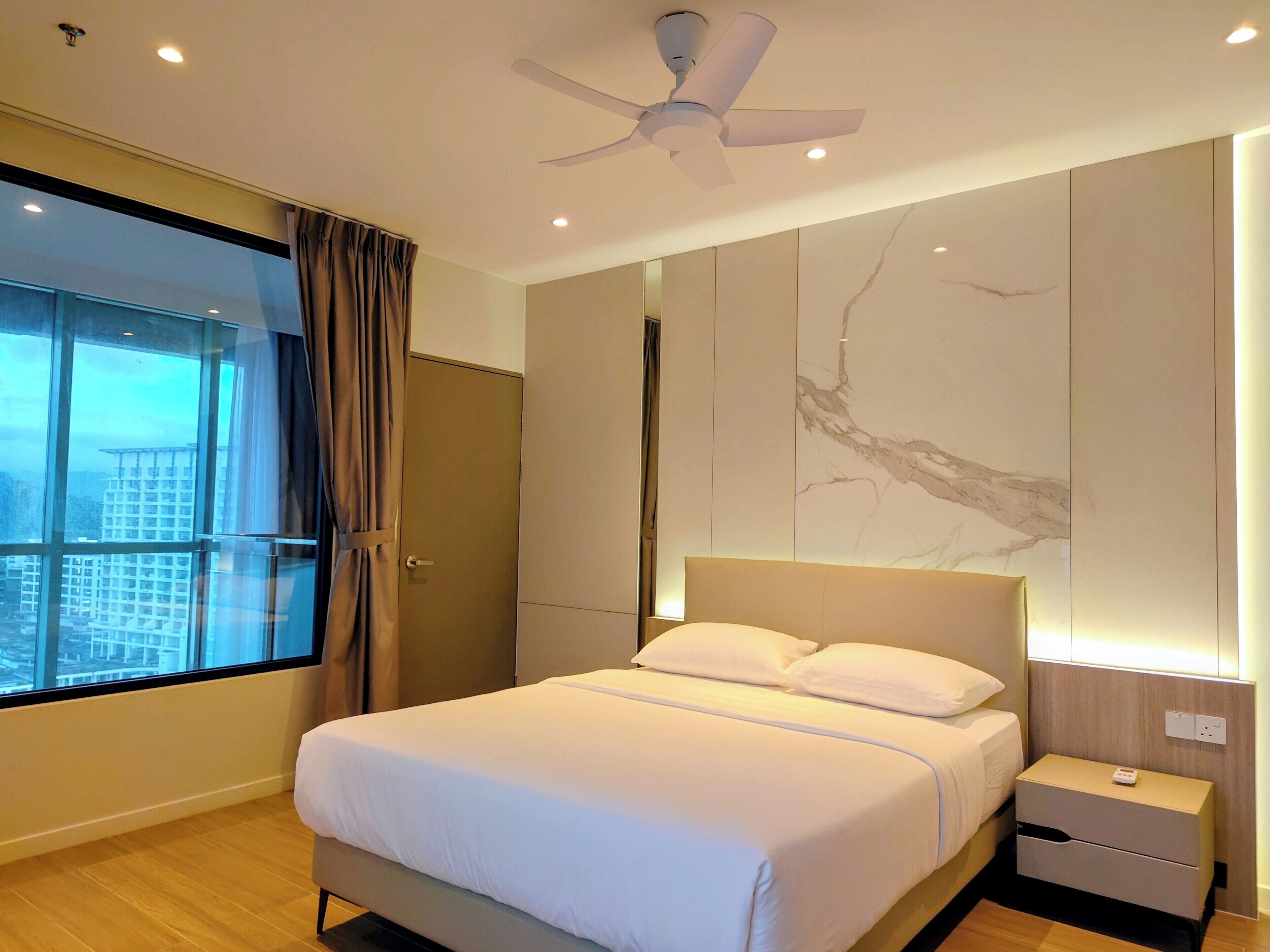 Een moderne hotelkamer met een groot bed, een plafondventilator, een nachtkastje en uitzicht op een stadsgezicht door een groot raam.