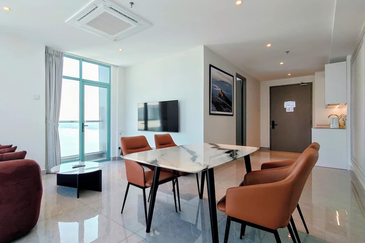 A modern hotel room with a large window, a flat-screen TV, a dining table with chairs, a red sofa, and a small side table.