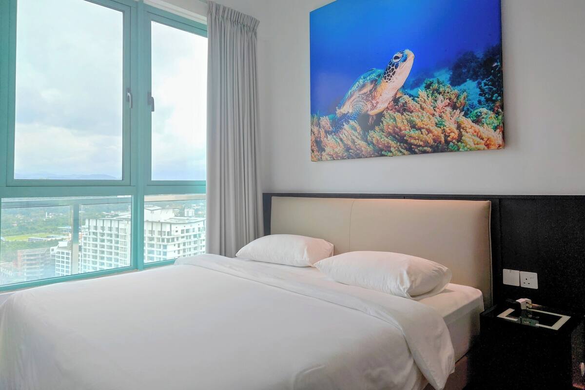 A hotel room with a large bed, a painting of a sea turtle, a window with a city view, and a nightstand with a lamp.