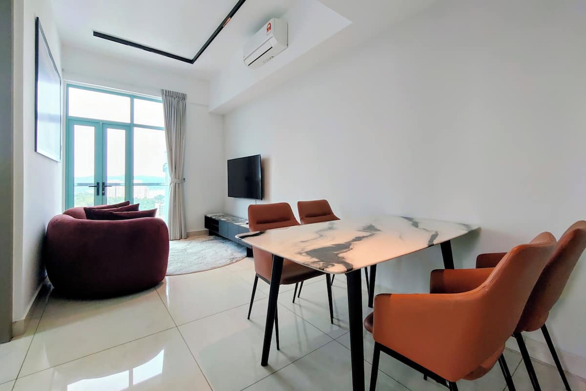 A modern hotel room with a large window, a flat-screen TV, a marble-top dining table, and orange chairs.
