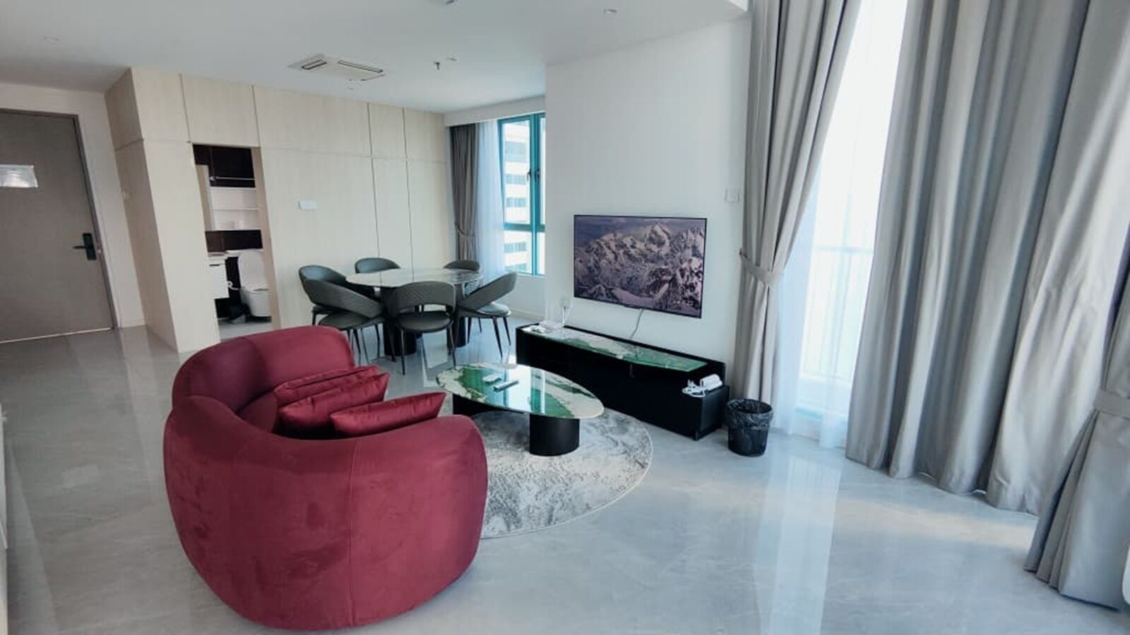 A modern living room with a red sofa, a glass coffee table, a dining area with chairs, a black entertainment unit, and a large window with curtains.