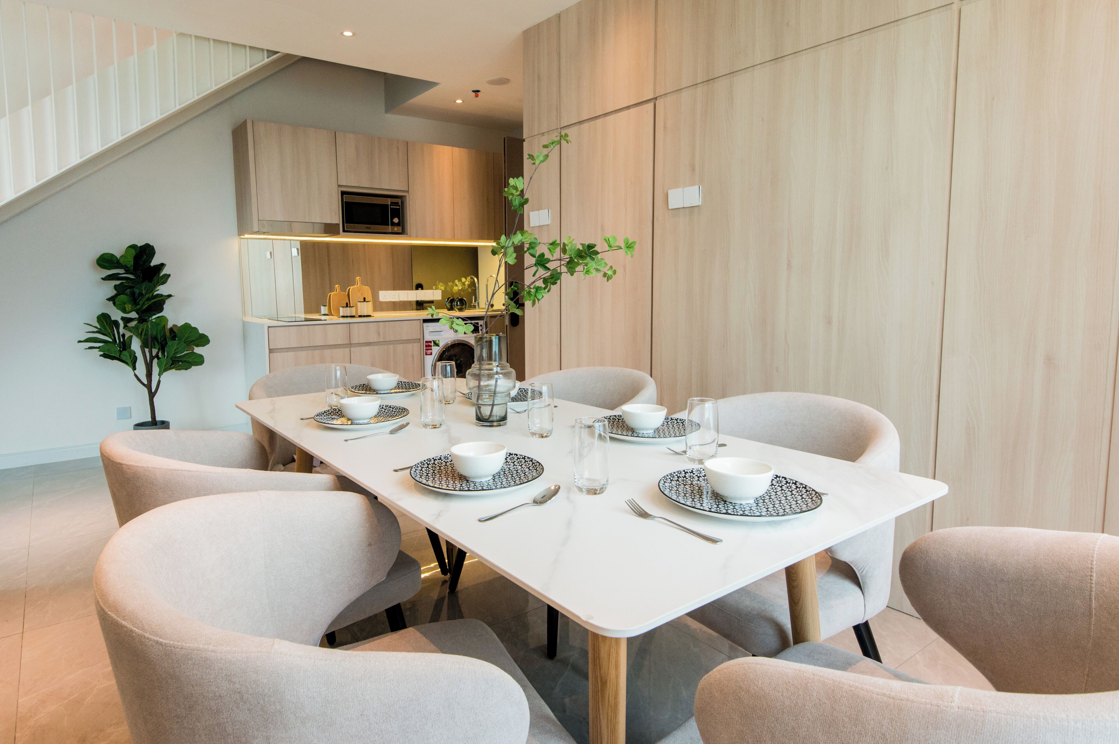 Un coin repas moderne avec une table blanche dressée pour six personnes, entourée de chaises de couleur beige. Une plante est placée sur le côté gauche de la table.