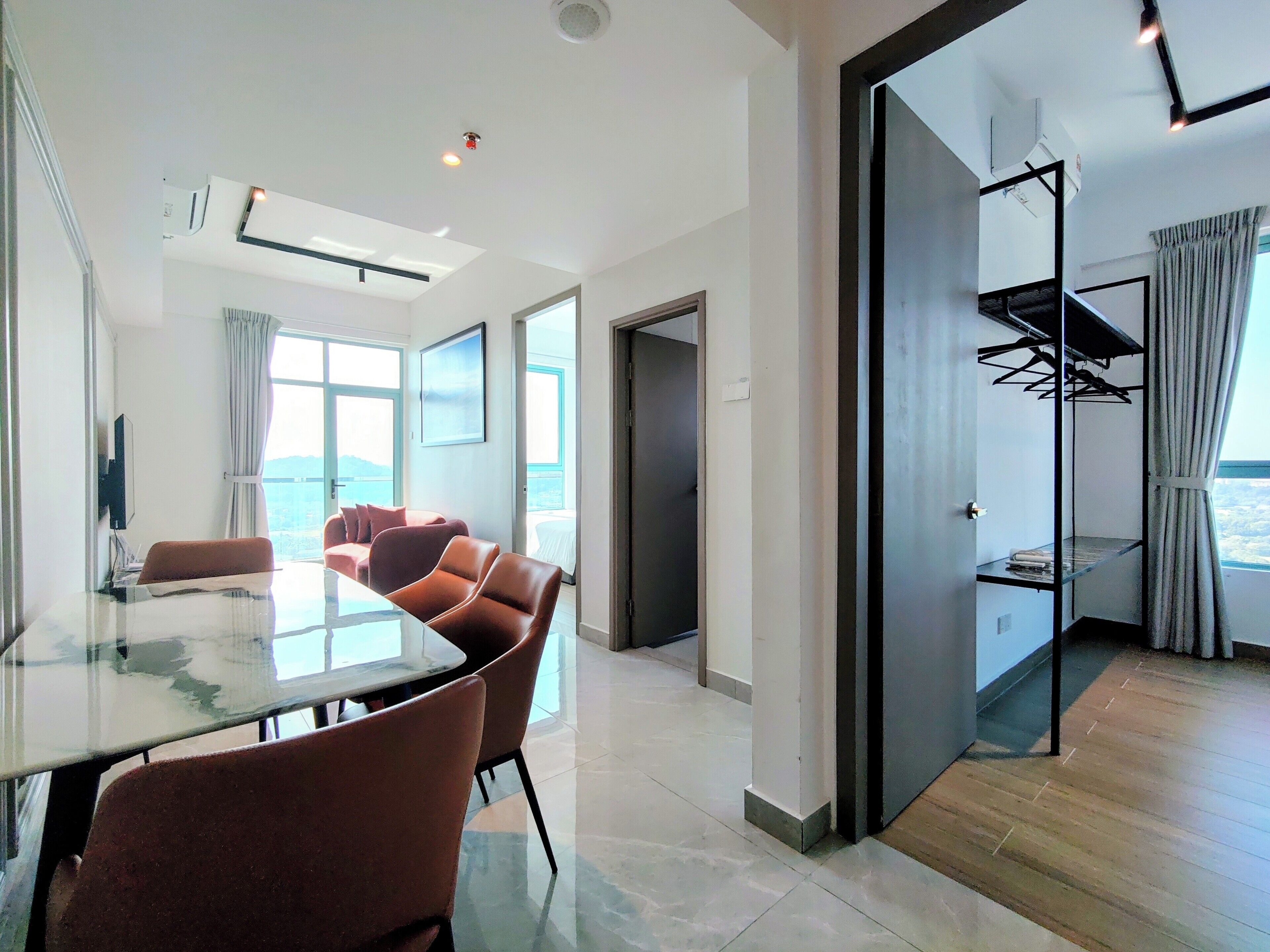 A modern hotel room with a glass dining table, leather chairs, a large window with a view, and a walk-in closet with a hanging rack.
