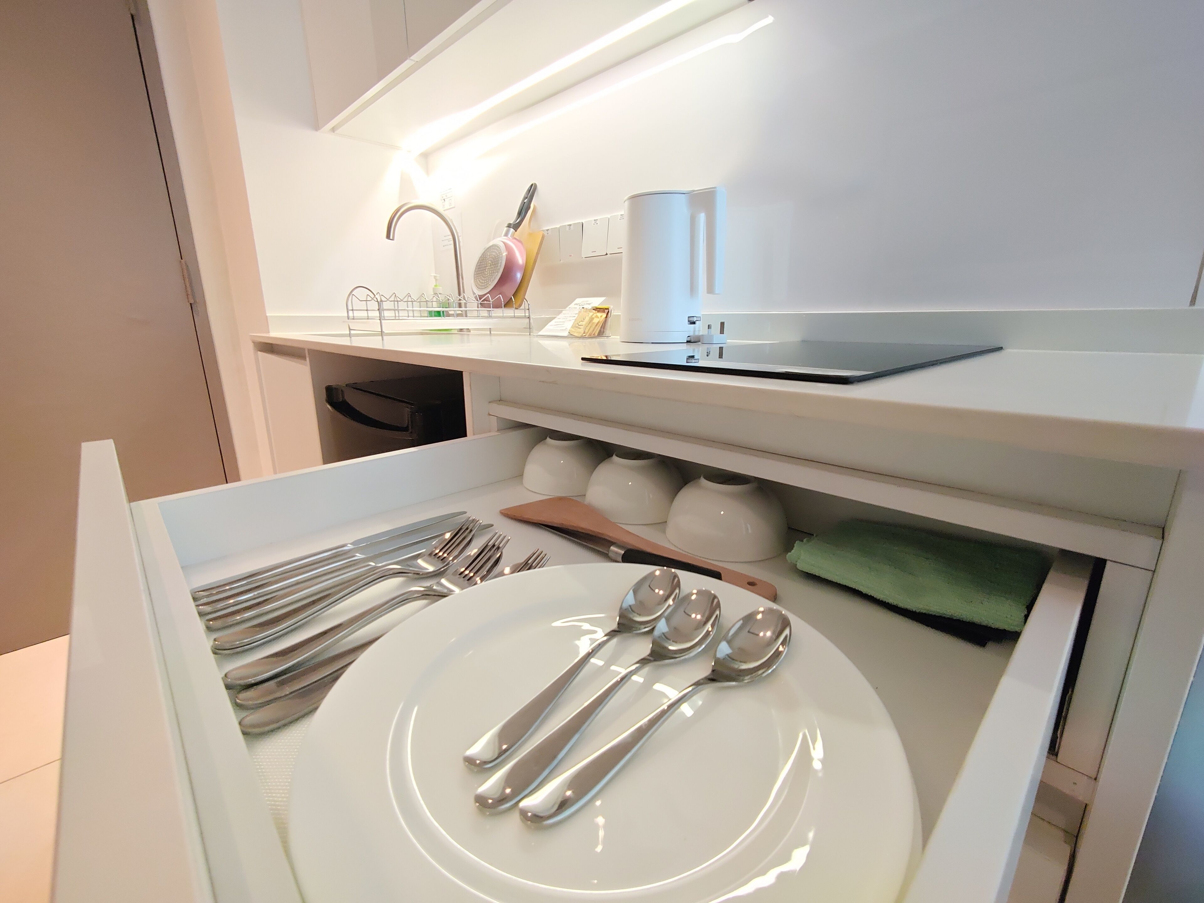 Une cuisine moderne avec des meubles blancs, un four encastré et un plan de travail sur lequel se trouvent une bouilloire, une machine à café et un ordinateur portable.